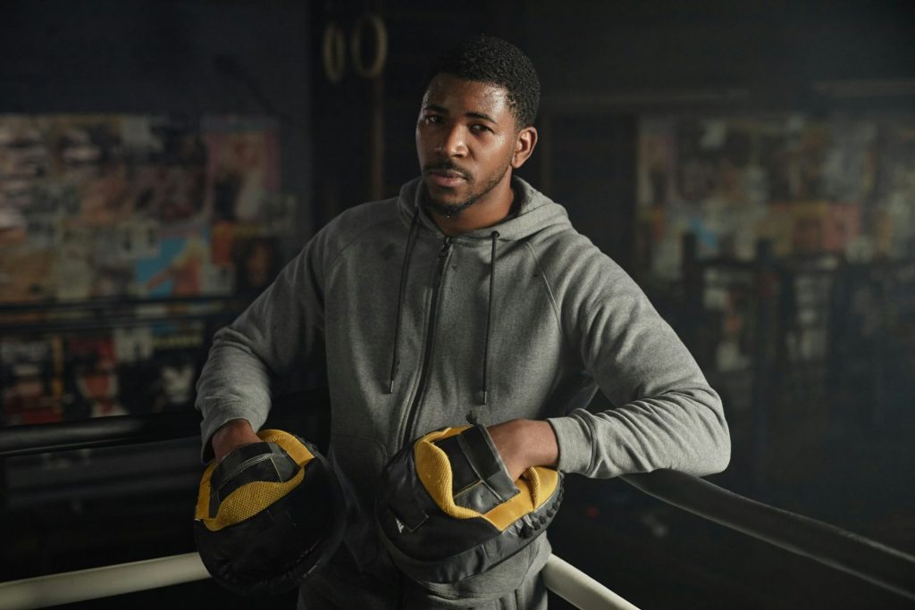 Young African American male boxing trainer in hoodie and pads