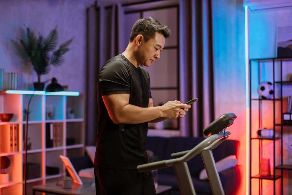 Portrait of active asian man in sportswear using smartphone, running using treadmill at home